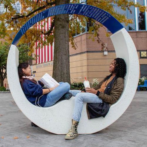 Two students seated on a sculptural bench outside on the Kean campus